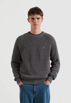 Grauer gestrickter Pullover mit Rippstruktur, Rundhalsausschnitt und subtilen Sprenkeln. Kombiniert mit blauen Jeanshosen, Hände in den Taschen.