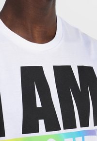 T-shirt en coton blanc avec un texte noir audacieux "I AM" et des accents dégradés colorés en dessous. Col rond, manches courtes.