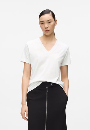 Donna con capelli scuri, indossa una maglietta bianca a scollo a V infilata in una gonna nera con cerniera frontale, in piedi con una mano in tasca davanti a uno sfondo bianco.