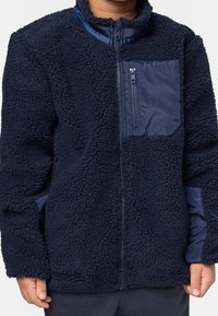 Enfant portant une veste polaire bleu marine avec une fermeture éclair complète et une poche poitrine zippée, debout devant un fond blanc uni.