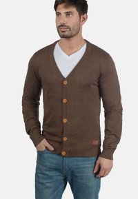 Brun strikket cardigan med V-hals, knappelås og ribbestrikket kant. Har fem treskknapper og bæres over en hvit T-skjorte.