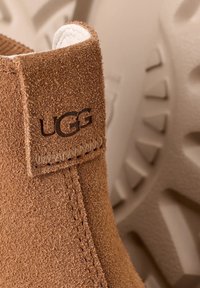 Stivale alla caviglia in suede marrone con una soffice fodera bianca, caratterizzato da una cucitura laterale e un logo UGG impresso sulla parte superiore. Suola testurizzata sullo sfondo.