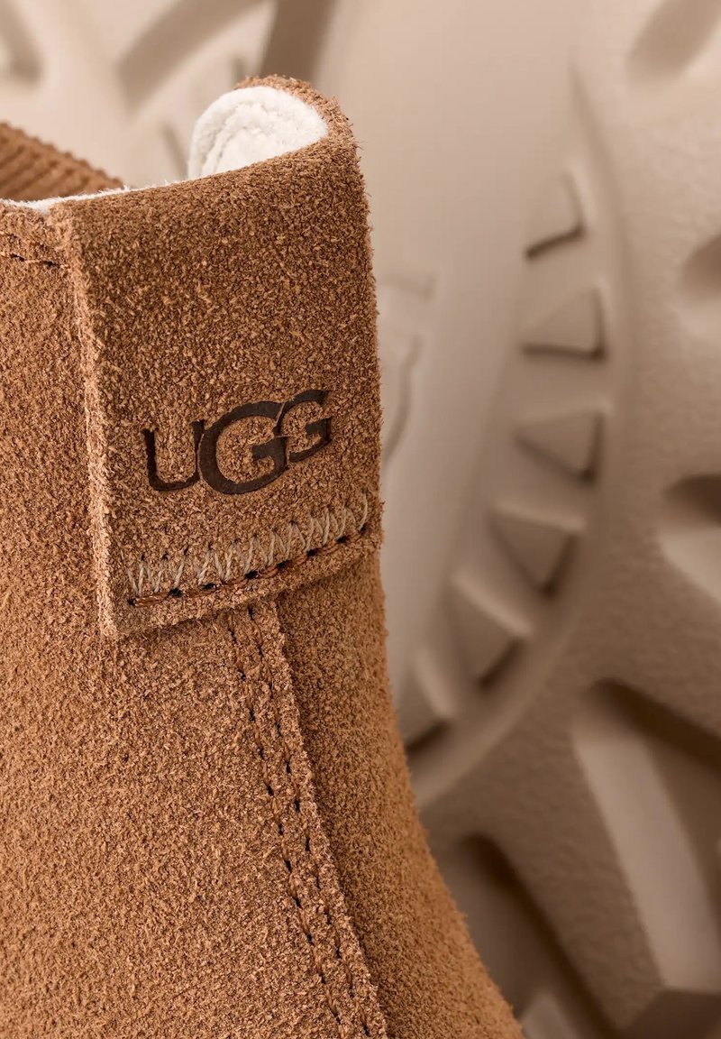 Brune ruskindsankelstøvle med en blød hvid foring, med en syet sidesøm og et UGG-logo prægnet på overdelen. Fodsål med tekstur i baggrunden.