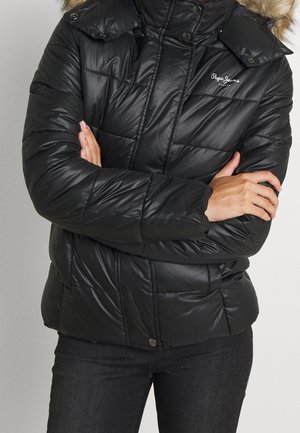Persoon draagt een zwarte pufferjas met een capuchon met bontkraag, gekruiste armen en een donkere broek, met het Pepe Jeans-logo op de borst zichtbaar.