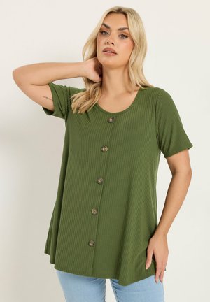 Donna bionda con indosso una maglia verde a maniche corte con bottoni e jeans azzurri, che posa con una mano dietro il collo.