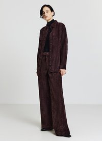 Giacca in velluto a coste bordeaux con grandi bottoni, abbinata a pantaloni a gamba larga dello stesso colore. Dolcevita nero sotto, scarpe nere eleganti.