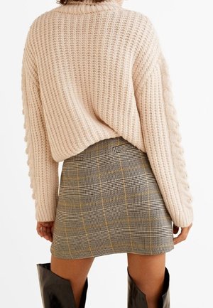 Femme portant un pull en maille épaisse beige, une mini-jupe à carreaux avec des touches jaunes, et des bottes noires montant jusqu'aux genoux, vue de dos.