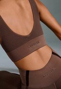 Brown ribbed sports bra with a low back, featuring textured panels and a contrasting band. Subtle logo detail at the waistband.