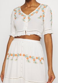 Blusa bianca corta con ricami floreali arancioni, maniche corte a campana e chiusura a bottoni. Abbinata a una gonna ricamata coordinata.