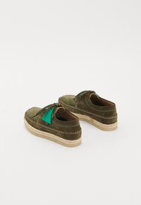 Des chaussures mocassins en daim vert olive avec des semelles en corde tressée et des étiquettes vertes, présentées sur un fond blanc.