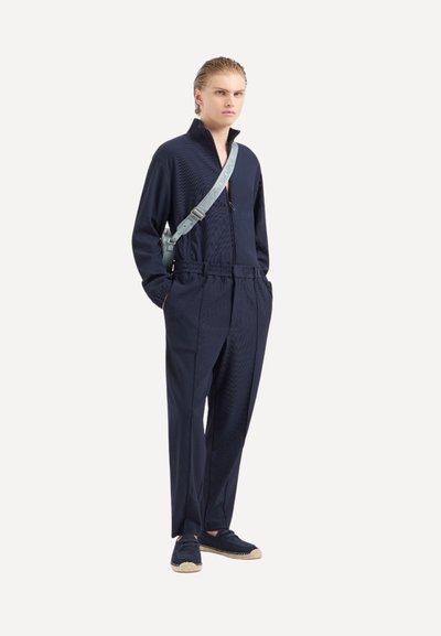 Tuta blu navy in tessuto texturizzato, con colletto alto, chiusura con zip e vita elastica. Abbinato a scarpe blu navy e una leggera tracolla.