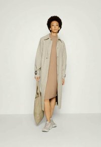 Trench coat xadrez longo em bege e castanho sobre um vestido justo castanho. Ténis cinzentos e uma grande bolsa tote bege completam o look.