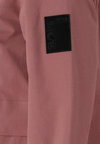 Rosa Jacke mit einem dezenten Nadelstreifenmuster. Verfügt über ein schwarzes, rechteckiges Stoffabzeichen mit der Aufschrift "SLOPE" am Oberarm. Insgesamt glatte Textur.