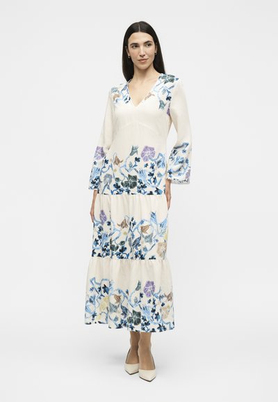Femme portant une longue robe blanche avec broderies florales bleues, violettes et beiges, manches évasées et talons blancs pointus, debout sur fond blanc.
