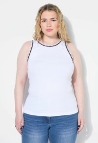 Witte mouwloze top met zwarte bies langs de halslijn en armsgaten, gemaakt van een geribbelde stof. Gepaard met lichtblauwe jeans.