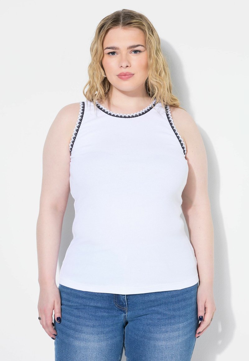 Witte mouwloze top met zwarte bies langs de halslijn en armsgaten, gemaakt van een geribbelde stof. Gepaard met lichtblauwe jeans.
