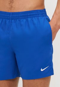 Blå badeshorts med elastisk linning, med et hvitt Nike swoosh-logo nederst til høyre og glatt, lett stoff.