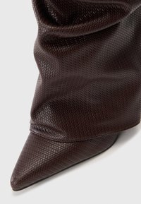 Botín de punta afilada de color marrón oscuro con un patrón texturizado en forma de escamas y detalle de parte superior arrugada sobre un fondo claro.