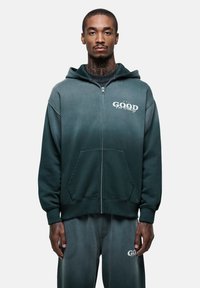 Felpa con cappuccio e zip in tessuto verde sfumato. Presenta tasche frontali, polsini a coste e il logo "GOOD" con dettagli bianchi.