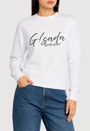 Frau trägt weißen Pullover mit schwarzem "G! sada Switzerland" Text, kombiniert mit blauen Jeans, Hand in der Tasche, neutraler Hintergrund.