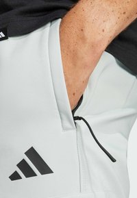 Lichtgrijze sport shorts met een zwart ritsvak, elastische tailleband en zwart Adidas-logo op het linkerpijpen. Gestructureerde stof.