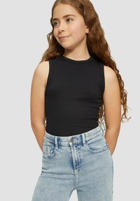 Meisje met lange, krullende haren in een mouwloze zwarte ribbed top die in hoge lichtblauwe denim jeans is gestopt, staand met de handen achter de rug.