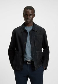 Zwarte suede jas met een kraag, voorzakken en knoopsluiting, gecombineerd met een lichtblauwe gestructureerde trui. Minimalistisch, modern ontwerp.