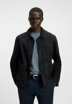 Veste en suédine noire avec un col, des poches avant et une fermeture à boutons, associée à un pull texturé bleu clair. Design minimaliste et moderne.