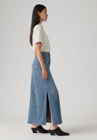 Šviesiai mėlyna džinsinė maksi sijonas su aukštu plyšiu ir tradiciniais siuvinėjimais; derinamas su trumpomis rankovėmis ir apykakle palaidine bei juodais aukštakulniais šlepečiais.