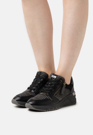 Jambes portant des baskets à lacets noires et grises avec fermeture éclair latérale et détails en tissu à carreaux sur fond blanc.