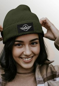 Olive green knit beanie with a black patch featuring a star and wings design. The hat has a textured finish and a cuffed edge.