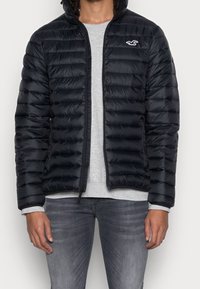 Black puffer jacket with horizontal quilted texture, zip closure, and a small logo on the left chest; paired with a gray long-sleeve shirt.