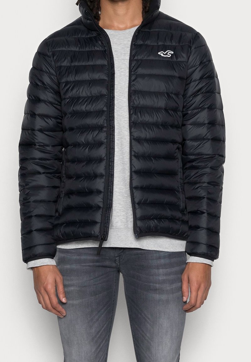 Black puffer jacket with horizontal quilted texture, zip closure, and a small logo on the left chest; paired with a gray long-sleeve shirt.