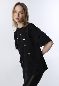 Czarny, luźny t-shirt z krótkim rękawem, ozdobiony okrągłymi, odbijającymi światło cekinami w różnych rozmiarach. Gładka faktura i swobodny krój.