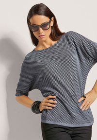 Grijze, getextureerde gebreide top met korte mouwen en een wijd uitgesneden halslijn. Gecombineerd met zwarte accessoires. Model poseert tegen een lichte achtergrond.