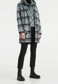 Abrigo de mezcla de lana gris con un patrón de cuadros en negro y azul, con grandes bolsillos, y combinado con una chaqueta interior acolchada verde y pantalones negros.