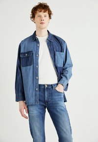 Camicia di denim in blu chiaro e scuro con due tasche frontali, chiusura a bottoni e vestibilità comoda, abbinata a jeans di denim blu medio.