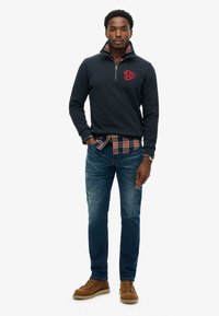 Pullover in pile blu navy con logo rosso, indossato sopra una camicia a quadri. Abbinato a jeans blu sbiaditi e stivali marroni con suole bianche.