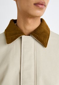 Chaqueta beige con un cuello de terciopelo marrón texturizado, que presenta un diseño limpio y un notable detalle de costura a lo largo de la abertura.