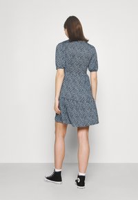 Modré květinové šaty s krátkými nafouknutými rukávy, povzneseným pasem a rozevlátou sukní. V kombinaci s černými vysokými teniskami. Minimalistické pozadí.