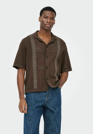 Man draagt een bruin kort mouw gebreid shirt met patroon van verticale strepen en blauwe spijkerbroek, staande met één hand in de zak.