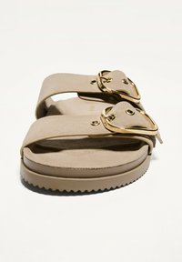 Béžové sandály se dvěma širokými pásky, zlatými přezkami a polstrovanou podrážkou. Podrážka má vroubkovaný okraj pro lepší uchopení. Měkká semišová textura.