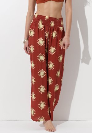 Donna con pantaloni a gamba larga color ruggine con motivi di sole gialli e un top corto marrone, in piedi a piedi nudi contro un muro bianco.