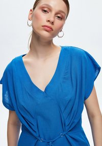 Blusa blu a maniche corte con scollatura a V profonda e vita cintrata, realizzata in tessuto morbido e testurizzato con dettagli arricciati, fissata con anelli.