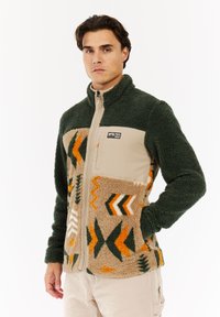 Mann trägt eine grüne und beige Fleecejacke mit orangefarbenen, wei�ßen und grünen geometrischen Mustern und steht mit einer Hand in der Tasche.