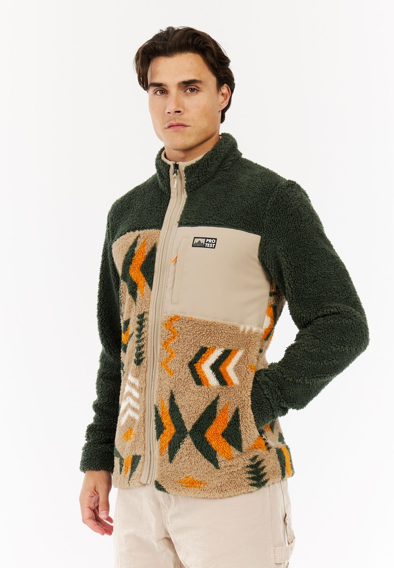 Mann trägt eine grüne und beige Fleecejacke mit orangefarbenen, weißen und grünen geometrischen Mustern und steht mit einer Hand in der Tasche.