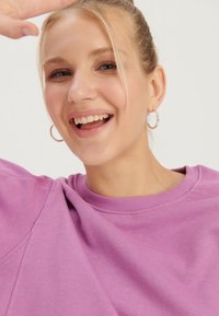 Jeune femme souriante aux cheveux blonds en chignon porte des boucles d'oreilles créoles argentées et un sweat-shirt violet, levant un bras près de son front.