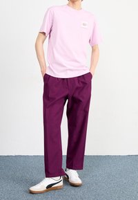 Osoba ubrana w jasnofioletowy t-shirt z napisem, ciemnofioletowe spodnie, białe sneakersy z czarnym paskiem, stojąca z rękami w kieszeniach na niebieskim dywanie.