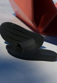 Black slide sandals made of rubber, featuring a wide strap and textured sole with ridges for grip. Brand logo visible on the side.