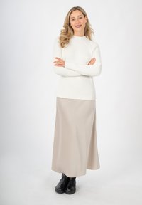 Weißes geripptes Strickpullover, beiger satinierter Maxirock und schwarze klobige Stiefeletten. Das Modell steht mit verschränkten Armen vor einem schlichten Hintergrund.